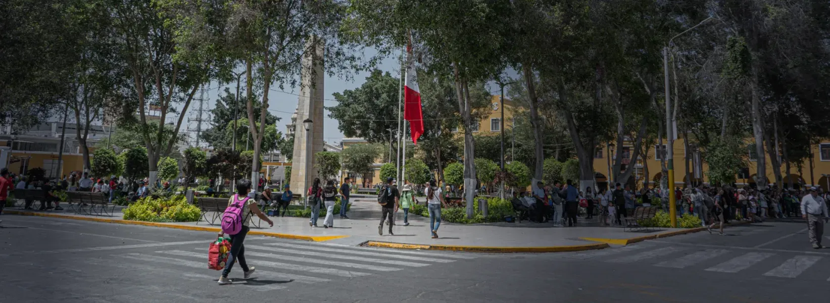 Parque con personas caminando, monumento, bandera del Perú y diversa vegetación y arboles visibles