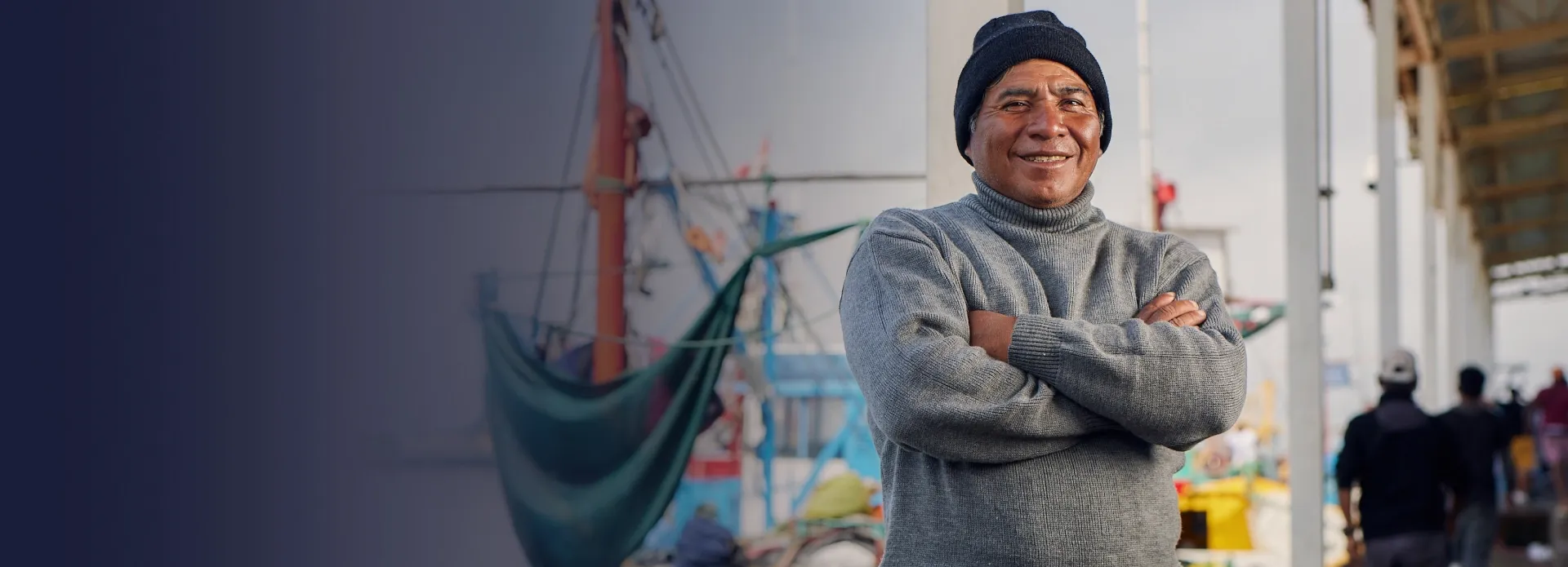 Pescador sonriendo cruzado de brazos, se ven botes y gente en el fondo desenfocados